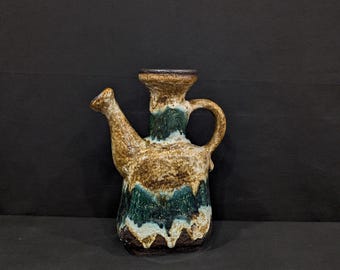 May include: A ceramic watering can with a spout and handle. The can has a brown, green, and tan glaze. The design has a textured appearance, with the colours blending together. The watering can is on a black surface.