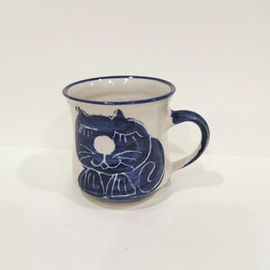 Può includere: Tazza in ceramica color crema con un disegno di gatto blu. Il gatto è raffigurato in uno stile stravagante e a cartoni animati. Il manico e il bordo della tazza sono anch'essi blu. Un'aggiunta affascinante e unica a qualsiasi cucina.