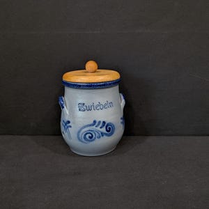 Pode incluir: Um pequeno pote de cerâmica azul claro com tampa de madeira e um pequeno botão de madeira. O pote tem redemoinhos decorativos azuis e a palavra "Zwiebeln" em escrita azul. Duas pequenas alças nas laterais.