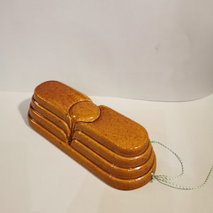 May include: A speckled, golden-brown ceramic soap dish. The rectangular dish features a raised, rounded section and tiered ridges. A green and white string is attached. The dish is approximately 18 cm long.