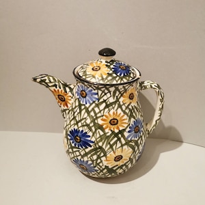 Op de afbeelding: Een keramische theepot met deksel, met een bloemmotief. De theepot is versierd met blauwe, gele en oranje madeliefachtige bloemen, tegen een achtergrond van groene grasachtige lijnen.