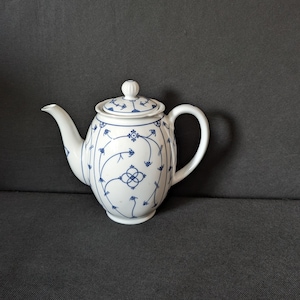 Può includere: Una teiera in porcellana bianca con un delicato motivo floreale blu composto da sottili steli curvi e piccoli fiori. La teiera include un coperchio abbinato con un piccolo pomello, un beccuccio curvo e un manico arrotondato sul lato.