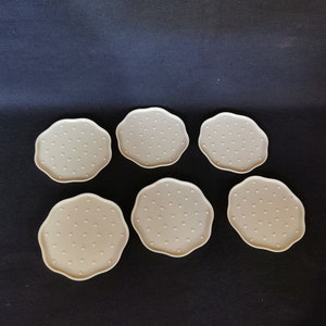 May include: Six beige coasters with a dotted pattern. The coasters are round and have a scalloped edge.