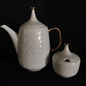 May include: White porcelain teapot and sugar bowl with gold accents and an intricate embossed pattern. The teapot has a tall, pointed lid and a gold handle. The sugar bowl has a lid with a small hole in the top.