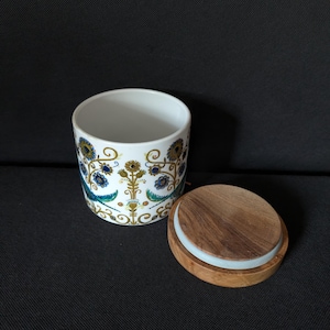 Puede incluir: Un tarro de cerámica blanco con un estampado floral en azul, dorado y turquesa, y una tapa de madera. El tarro es cilíndrico y abierto, con la tapa colocada al lado. El fondo es de tela gris oscuro.