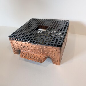 Puede incluir: Una caja metálica de color cobre con una superficie texturizada y una parte superior negra en forma de rejilla. La caja tiene una abertura cuadrada en el centro de la parte superior y está elevada por pequeños pies. La superficie de la caja tiene un patrón en relieve de formas ovaladas.