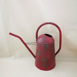 Könnte beinhalten: Eine rote Gießkanne aus Metall mit gebogenem Griff und langem Ausguss. Die Kanne hat eine Used-Optik, mit Bereichen abgenutzter Farbe, die das darunter liegende Metall freilegen. Die Gießkanne ist für den Gartenbau konzipiert und hat eine klassische, Vintage-Ästhetik.