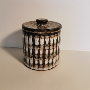 Op de afbeelding: Een bruine en witte keramische pot met een deksel. De pot heeft een geometrisch patroon van witte vierkanten en bruine lijnen.