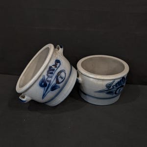 Pode incluir: Uma caneca de cerâmica branca com uma alça preta e o texto "But First, Coffee" em preto, com uma fonte manuscrita. A caneca está sobre uma superfície branca.