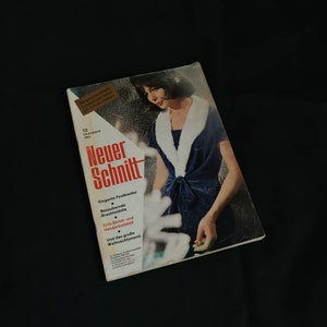 Op de afbeelding: Een tijdschriftcover met een vrouw in een blauwe jurk met een witte bontkraag. Het tijdschrift heet "Neuer Schnitt" en de uitgave is 12 december. De cover bevat ook de tekst "Elegante Festkleider, Bezaubernde Brautmodelle, 115A-Strick-und Handarbeitstell, Und das große Weihnachtsmenü."