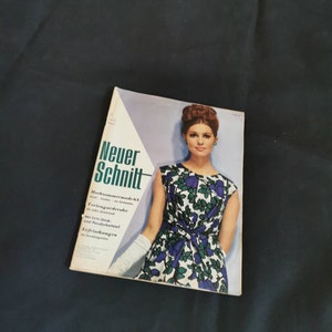 Op de afbeelding: Een vintage tijdschriftcover met een vrouw die een blauw-groene bloemenjurk draagt. Het tijdschrift is getiteld "Neuer Schnitt" en heeft de tekst "Hochsommermode 63" en "Ferien-Garderobe" op de omslag.