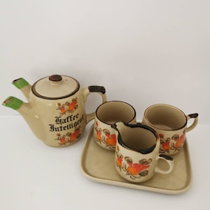 Puede incluir: Un juego de té de cerámica vintage con una tetera, tres tazas y una jarra de crema. La tetera tiene un pico y un asa verdes y presenta el texto "Kaffee Intelligent". Las tazas y la jarra de crema están decoradas con diseños florales de color naranja y marrón.