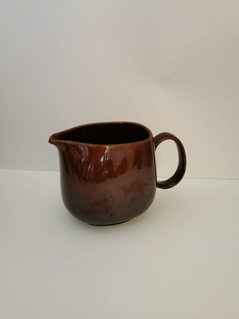 May include: A brown ceramic creamer pitcher with a rounded body and a handle. The pitcher is glazed with a glossy finish.