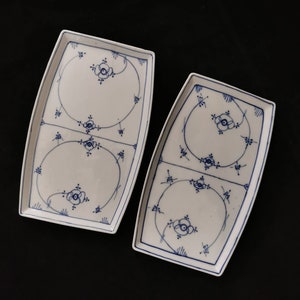 Puede incluir: Dos fuentes rectangulares blancas con diseños florales y lineales azules. Cada fuente tiene una forma rectangular redondeada con un borde azul y un motivo floral central. Las fuentes están dispuestas sobre una superficie negra.