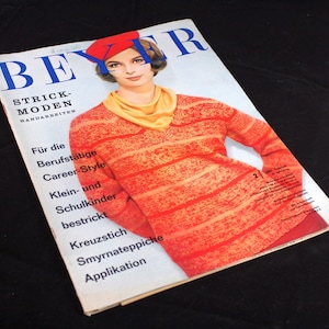 Pode incluir: Uma capa de revista com uma mulher vestindo um suéter listrado vermelho e laranja. A revista é intitulada "Beyer Strick-Moden Handarbeiten" e tem texto sobre estilo de carreira, tricô para crianças, ponto cruz e tapetes Smyrna.