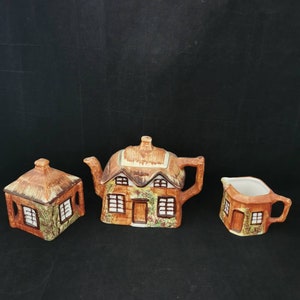 Könnte beinhalten: Ein Cottage-Tee-Set mit Teekanne, Zuckerdose und Milchkännchen. Die Keramikstücke sind mit brauner, grüner und weißer Farbe verziert, um einem kleinen Haus mit Fenstern und einem Strohdach zu ähneln.