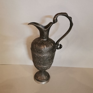 May include: An antique silver-coloured jug with an ornate floral design. The jug has a tall, slender body, a curved handle, and a spout. The base is round and elevated. The jug is likely made of metal and is suitable for decorative purposes.