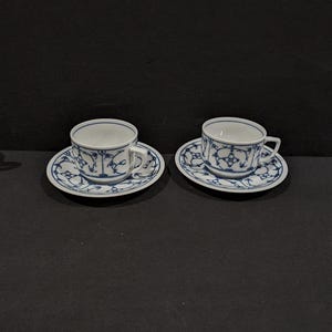 Könnte beinhalten: Zwei weiße Teetassen und Untertassen mit blauem Blumenmuster. Die Tassen haben kleine Henkel und stehen auf passenden Untertassen. Das blaue Design umfasst florale und geometrische Elemente und erzeugt einen klassischen, eleganten Look.