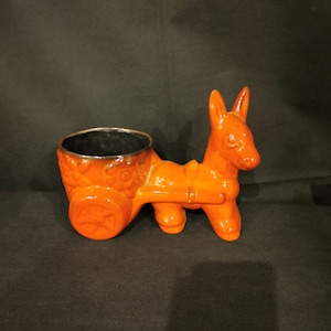 Puede incluir: Maceta de cerámica naranja en forma de burro con un carro de ruedas. El burro tiene un interior esmaltado negro.
