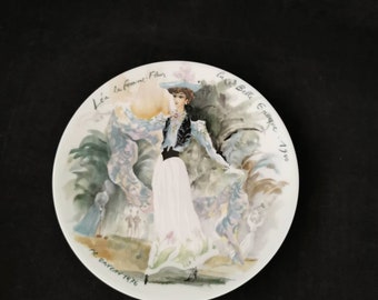 Pode incluir: Um prato de porcelana branca com uma pintura de aquarela de uma mulher com um vestido azul e branco. O prato é decorado com o texto "L'ea la femme-Fleur" e "La Belle Epoque - 1900" e a assinatura "P.G. Gaveau 1976".