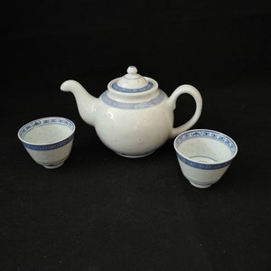Puede incluir: Una tetera blanca con un diseño floral azul y blanco y dos tazas de té a juego. La tetera tiene una tapa y un pico. Las tazas de té son pequeñas y tienen un borde.