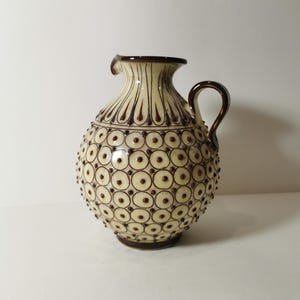 Puede incluir: Jarra de cerámica color crema con asa y borde marrón. El cuerpo está decorado con un patrón de círculos y puntos marrones. La jarra tiene forma redondeada y un pequeño pico.