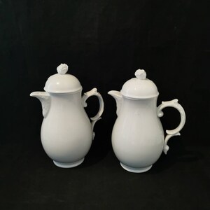 Op de afbeelding: Twee witte keramische koffiepotten met deksels, elk met een roosvormige knop. De potten hebben gebogen handvatten en tuiten, tegen een zwarte achtergrond. Het ontwerp is eenvoudig en elegant.