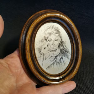 Könnte beinhalten: Ovaler Holzrahmen mit einer detaillierten Schwarz-Weiß-Illustration einer Mutter und eines Kindes. Das Kunstwerk ist vor einem silbernen Hintergrund platziert, und der Rahmen hat eine dunkelbraune Oberfläche.
