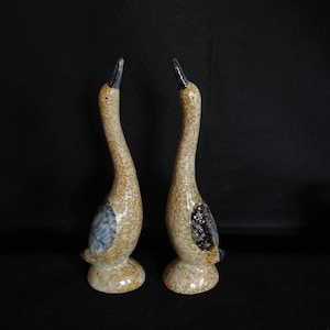 Puede incluir: Dos figuras de ganso de cerámica con cuerpos moteados de color canela, detalles en azul y negro y cuellos curvos. Los gansos tienen un acabado brillante y se exhiben sobre un fondo negro. Estas figuras decorativas son probablemente saleros y pimenteros.