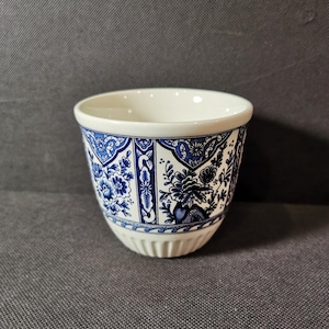 Może przedstawiać: Biały ceramiczny kubek z niebiesko-białym wzorem kwiatowym. Kubek ma zaokrąglone dno i rozszerzającą się krawędź.