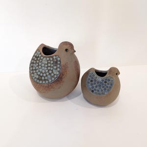 Puede incluir: Dos jarrones de cerámica con forma de pájaro. El jarrón más grande tiene un cuerpo marrón con un diseño de ala texturizado y moteado azul. El jarrón más pequeño refleja el diseño. Ambos tienen un interior oscuro y una forma de pájaro, adecuados para la decoración del hogar.