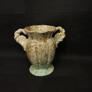 Könnte beinhalten: Kleine, dekorative Keramikvase mit einer strukturierten, gesprenkelten braun-grünen Glasur. Die Vase hat zwei verzierte Griffe und einen gewellten Rand. Sie steht auf einem kleinen, runden Sockel. Das Design erinnert an einen Vintage- oder Antik-Stil.