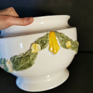 Può includere: Vaso in ceramica bianca con un design floreale in rilievo in tonalità di verde, giallo e rosa. Il vaso ha una base larga e rotonda e un bordo leggermente svasato.