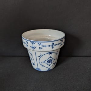 Könnte beinhalten: Ein weißer Blumentopf aus Keramik mit blauem Blumenmuster. Der Topf hat einen ausgestellten Rand und einen zylindrischen Körper. Das blaue Design umfasst stilisierte Blumen, Blätter und geometrische Formen. Der Topf ist leer und steht auf einer dunkelgrauen Oberfläche.