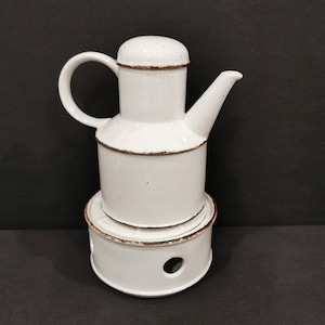 Op de afbeelding: Witte theepot van keramiek met een bruine rand en een bijpassende witte keramische warmhoudplaat. De theepot heeft een rond lichaam en een tuit aan de zijkant. De warmhoudplaat heeft een gat in het midden.