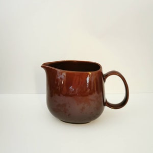May include: A brown ceramic pitcher with a rounded body and a handle. The pitcher has a glossy finish.