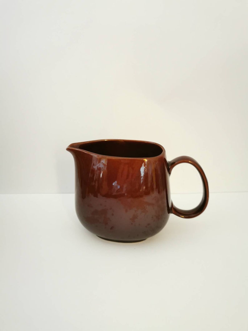 May include: A brown ceramic pitcher with a rounded body and a handle. The pitcher has a glossy finish.