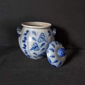 May include: A ceramic jar with a lid, featuring a blue and white floral design. The jar has two small handles and a rounded shape. The lid is placed next to the jar, showcasing the same blue floral pattern.