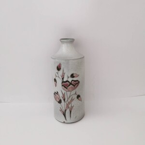 Könnte beinhalten: Eine weiße Keramikvase mit einem Blumendesign in Rosa- und Brauntönen. Die Vase hat eine zylindrische Form und einen schmalen Hals.