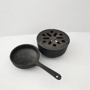 Könnte beinhalten: Eine schwarze gusseiserne Bratpfanne mit langem Griff und rundem Boden. Die Bratpfanne steht neben einem schwarzen gusseisernen Brenner mit kreisförmigem Design.