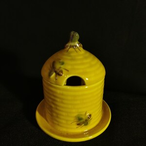 Może przedstawiać: Żółty ceramiczny słoik na miód w kształcie ula z małym otworem na górze. Słoik ma pasującą żółtą podstawkę i przedstawia namalowane pszczoły po bokach.