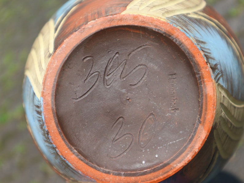 May include: The bottom of a brown ceramic vase with a blue and yellow design. The vase is marked with the text "325" and "Handarbeit".