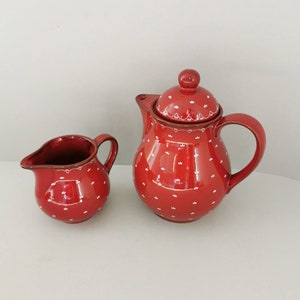 Puede incluir: Un juego de té de cerámica roja con una jarra de crema a juego. Ambas piezas están adornadas con patrones florales blancos y un borde blanco. La tetera tiene tapa y asa curva. El juego es probablemente para servir té o café.