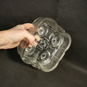 Könnte beinhalten: Eine quadratische Glasschale aus klarem Glas mit einem erhabenen, kreisförmigen Design. Die Schale hat einen leicht gewellten Rand.