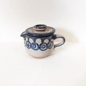 Peut inclure: Une théière en céramique avec un couvercle. La théière est principalement beige avec un bord et une anse bleu foncé. Le corps présente un motif répétitif de cercles bleus. Le couvercle est bleu foncé avec un bouton marron.
