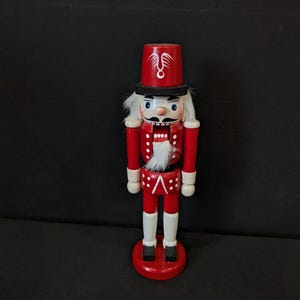 Könnte beinhalten: Eine rot-weiß lackierte Holz-Nussknacker-Figur. Die Figur hat einen roten Hut mit weißem Design, einen schwarzen Rand und weißes Haar. Der Nussknacker hat einen weißen Schnurrbart und Bart sowie eine rote Uniform mit weißen Akzenten.