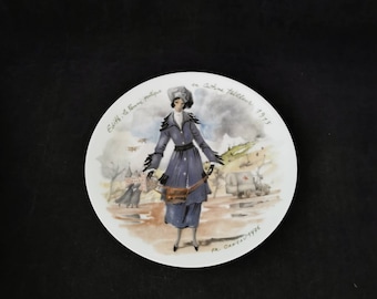 Puede incluir: Un plato de cerámica blanco con una ilustración colorida de una mujer con un abrigo y un sombrero azules caminando en un paisaje nevado. El texto "Edith - la Femme pratique en Costume Tailleur 1915" está escrito alrededor del borde del plato.