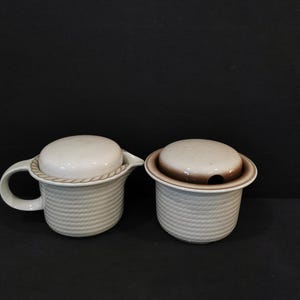 Puede incluir: Un juego de crema y azucarero de cerámica blanca con un patrón tejido y detalles marrones. La jarra tiene un asa y una tapa. El azucarero tiene una tapa con un pequeño agujero en la parte superior.