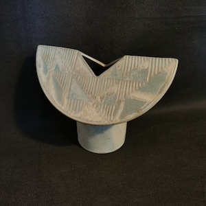 May include: A light blue ceramic vase with a unique, open design. The vase has a half-moon shape with a cut-out section and a textured surface. It stands on a short, cylindrical base. The vase is approximately 15 cm wide and 13 cm tall.