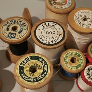 Puede incluir: Una colección de carretes de hilo de madera vintage con etiquetas en alemán. Los carretes son de varios colores y tienen diferentes etiquetas, incluyendo "MEZ" y "ACKERMANN'S".